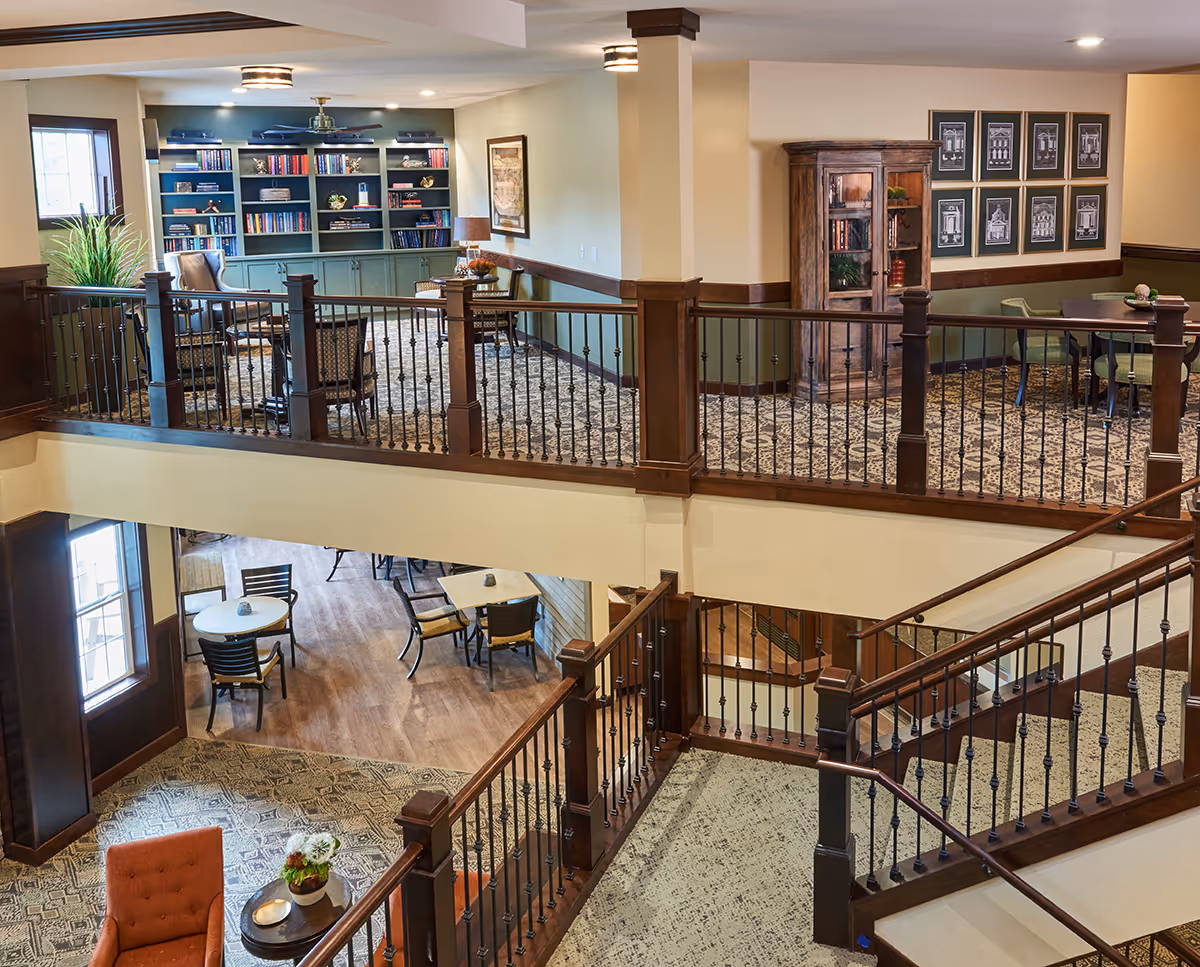 Interior view of a senior living facility showing a two-level common area with wooden railings and carpeted stairs. The upper level features a seating area with chairs and tables, a bookshelf filled with books, framed pictures on the wall, and a wooden cabinet. The lower level has round tables with chairs and large windows letting in natural light.