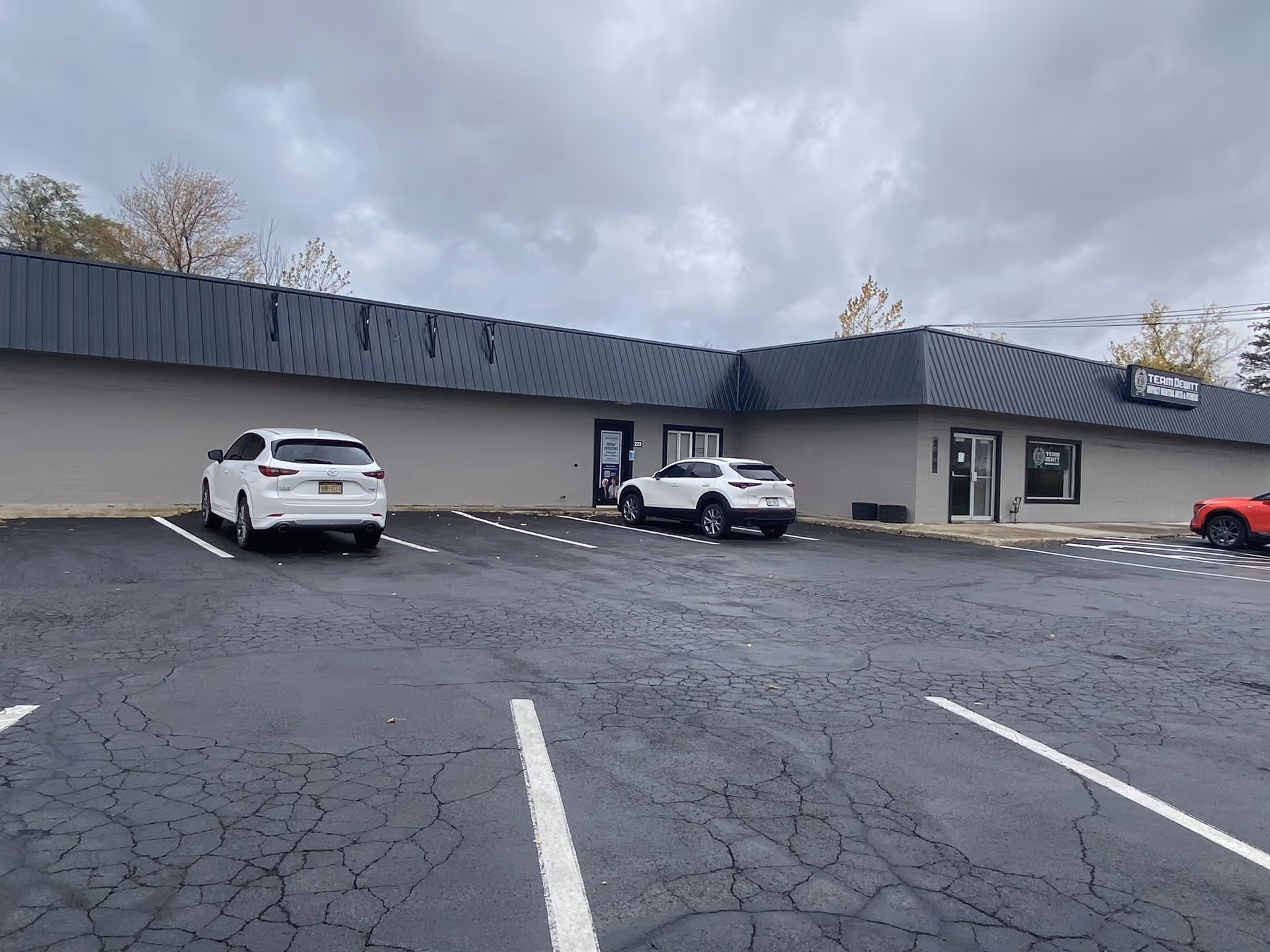 Long, low gray building with a metal roof and parked cars in a cracked lot.