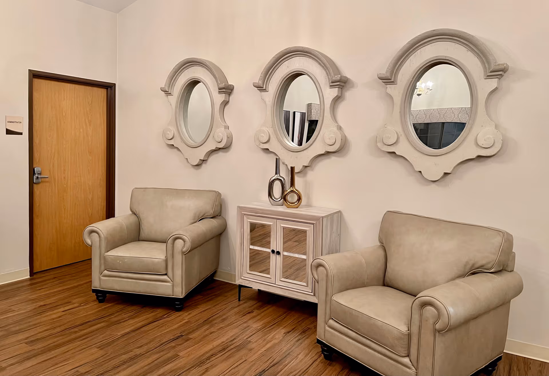 A cozy seating area with two beige armchairs placed on a wooden floor. Between the chairs is a small cabinet with glass doors and two decorative vases on top. Above the cabinet are three ornate oval mirrors mounted on a light-colored wall. To the left, there is a wooden door with a sign that reads 'Administrator'.