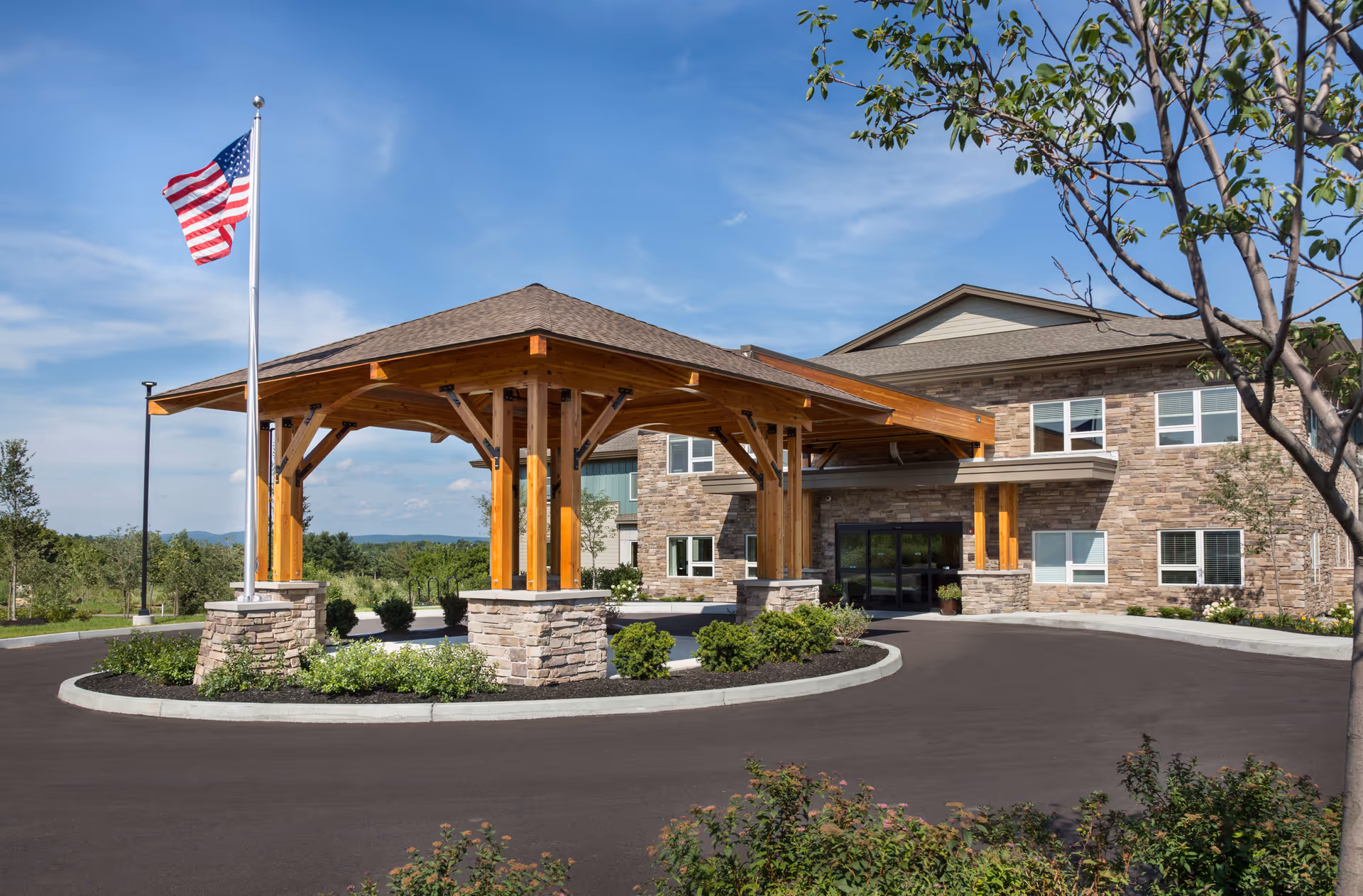 Exterior view of The Residence at Quarry Hill featuring a stone and wood entrance canopy with an American flag on a flagpole, surrounded by landscaped greenery and a clear blue sky.