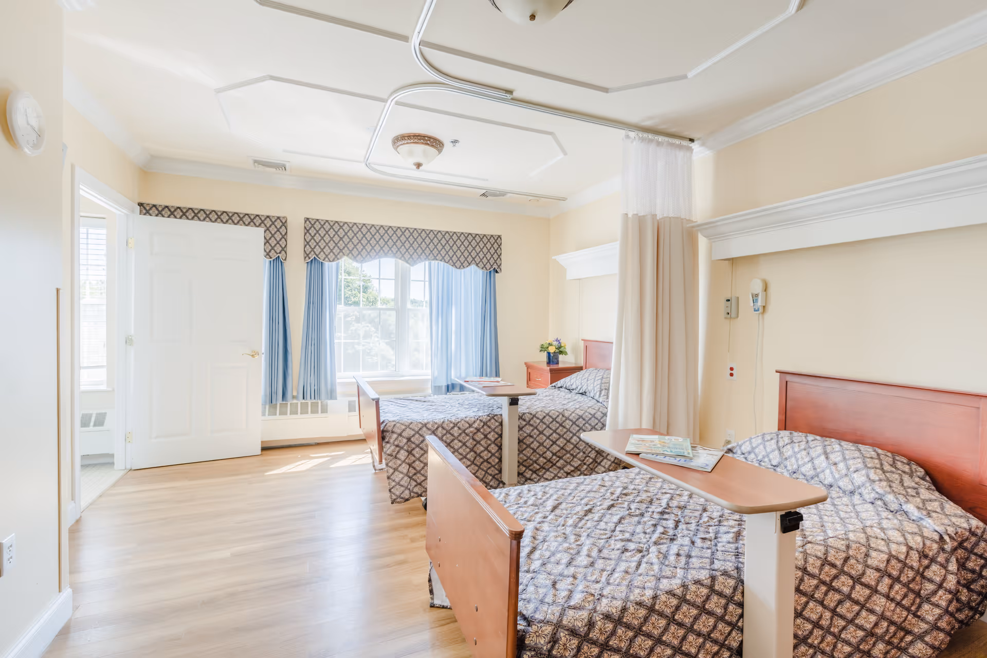 A bright and clean senior living bedroom with two hospital-style beds separated by a privacy curtain. Each bed has a patterned bedspread and an overbed table with magazines. The room features large windows with blue curtains and a decorative valance, light wood flooring, and cream-colored walls. There is a door open to an adjoining room or bathroom.