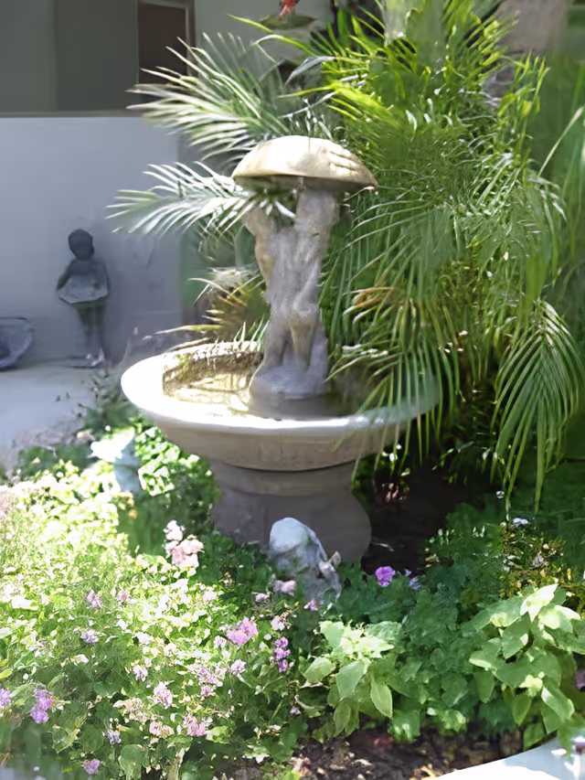 A stone garden fountain topped by a mushroom-shaped cap surrounded by palm fronds and blooming plants.