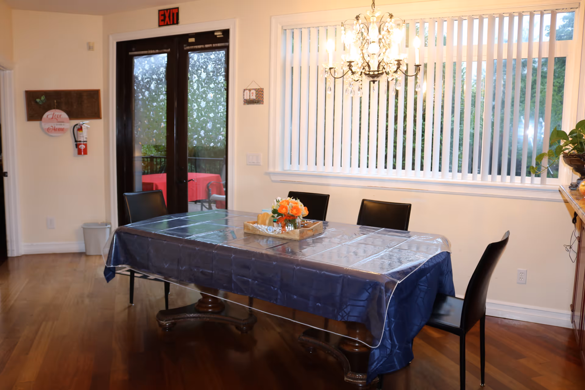 A dining area with a rectangular table covered by a blue tablecloth and a clear plastic cover. Four black chairs surround the table. A wooden tray with a small flower arrangement is placed in the center of the table. Behind the table is a large window with vertical blinds and a chandelier hanging from the ceiling. To the left, there are double glass doors with frosted floral patterns and an exit sign above them. A fire extinguisher and a decorative wall hanging are mounted on the wall near the doors.