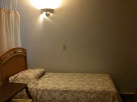 Single twin bed with patterned bedspread against a plain wall lit by a wall sconce in a small bedroom.