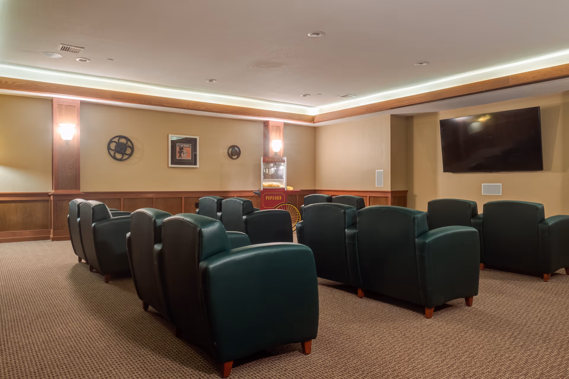A cozy senior living facility theater room with two rows of green leather armchairs facing a large flat-screen TV mounted on the wall. The room has warm beige walls with wooden paneling, soft lighting from wall sconces, and a popcorn machine in the corner.