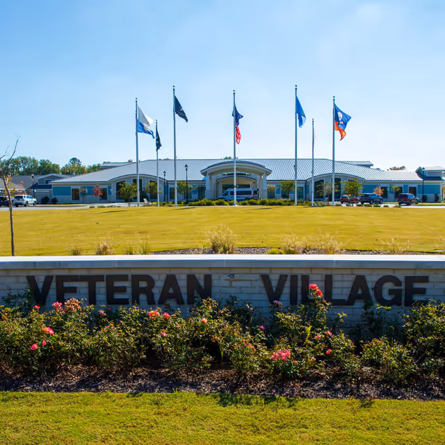 Front view of Veteran Village senior living facility with flags and a landscaped entrance sign.
