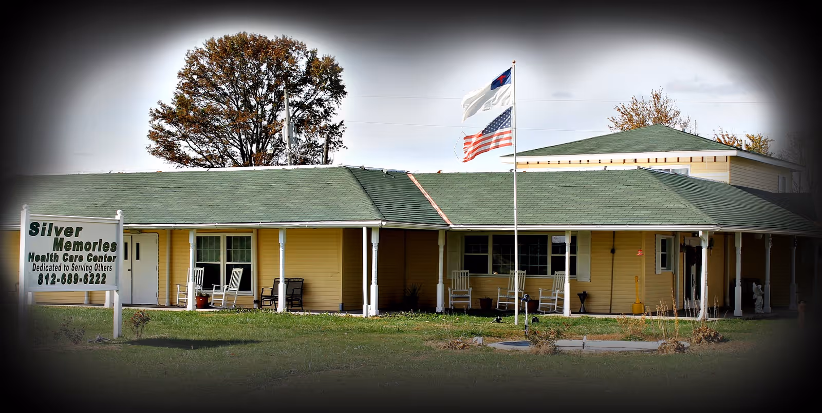 Exterior view of Silver Memories Health Care Center, a single-story yellow building with a green roof. There are rocking chairs on the covered porch and two flags flying on a flagpole in front of the building. A sign in the lawn displays the facility's name and phone number.