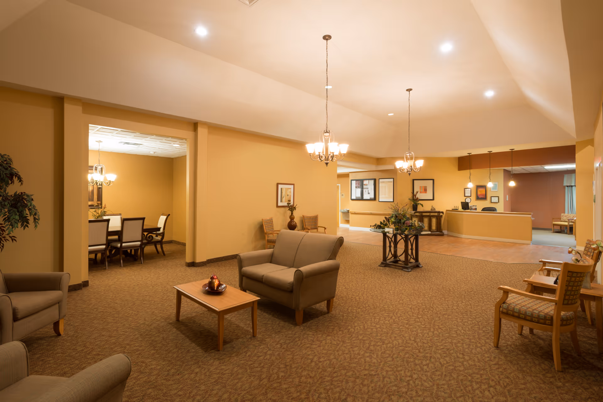 A spacious and warmly lit common area in a senior living facility with beige walls and carpeted floors. The room features comfortable seating including armchairs and a loveseat around a wooden coffee table. There are chandeliers hanging from the ceiling, a small table with a floral arrangement, and a reception desk in the background. To the left, there is an open doorway leading to a dining area with a table and chairs.