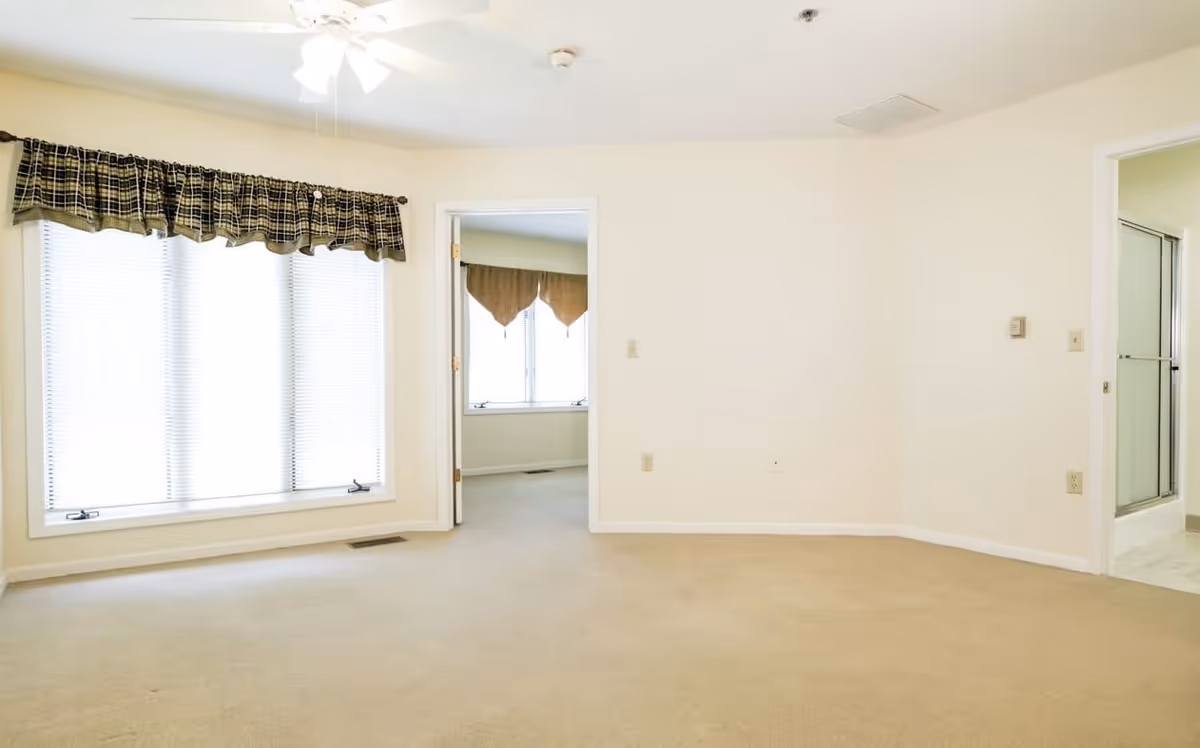 Empty room with beige carpet and cream-colored walls, a large window with blinds and a plaid valance, a ceiling fan with lights, and two doorways leading to another room and a bathroom with a glass shower door.