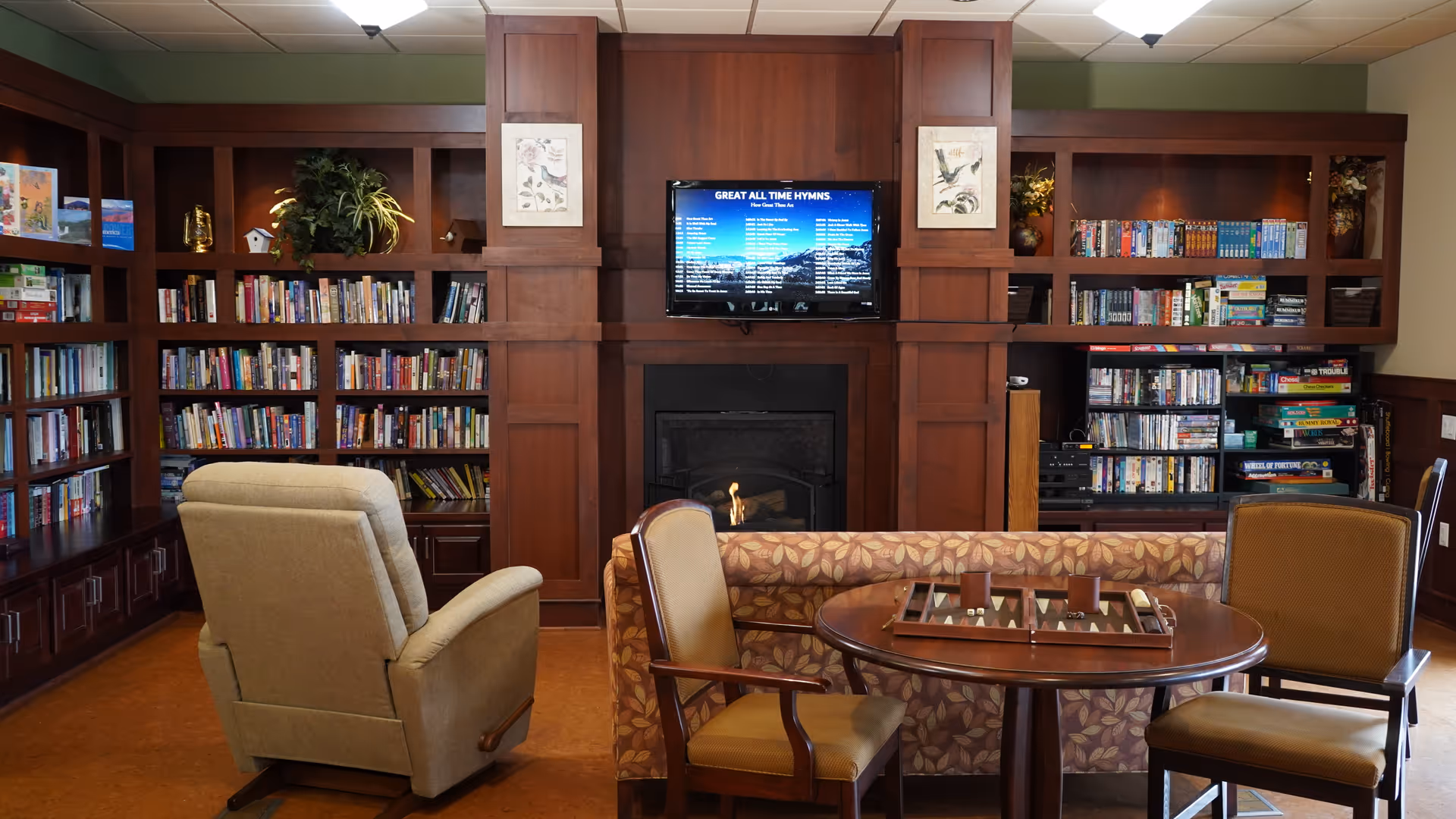 A cozy living room area with wooden bookshelves filled with books and board games. There is a beige recliner chair, a patterned sofa, and two wooden chairs around a round table with a backgammon game set on it. A flat-screen TV is mounted above a fireplace in the center of the room.