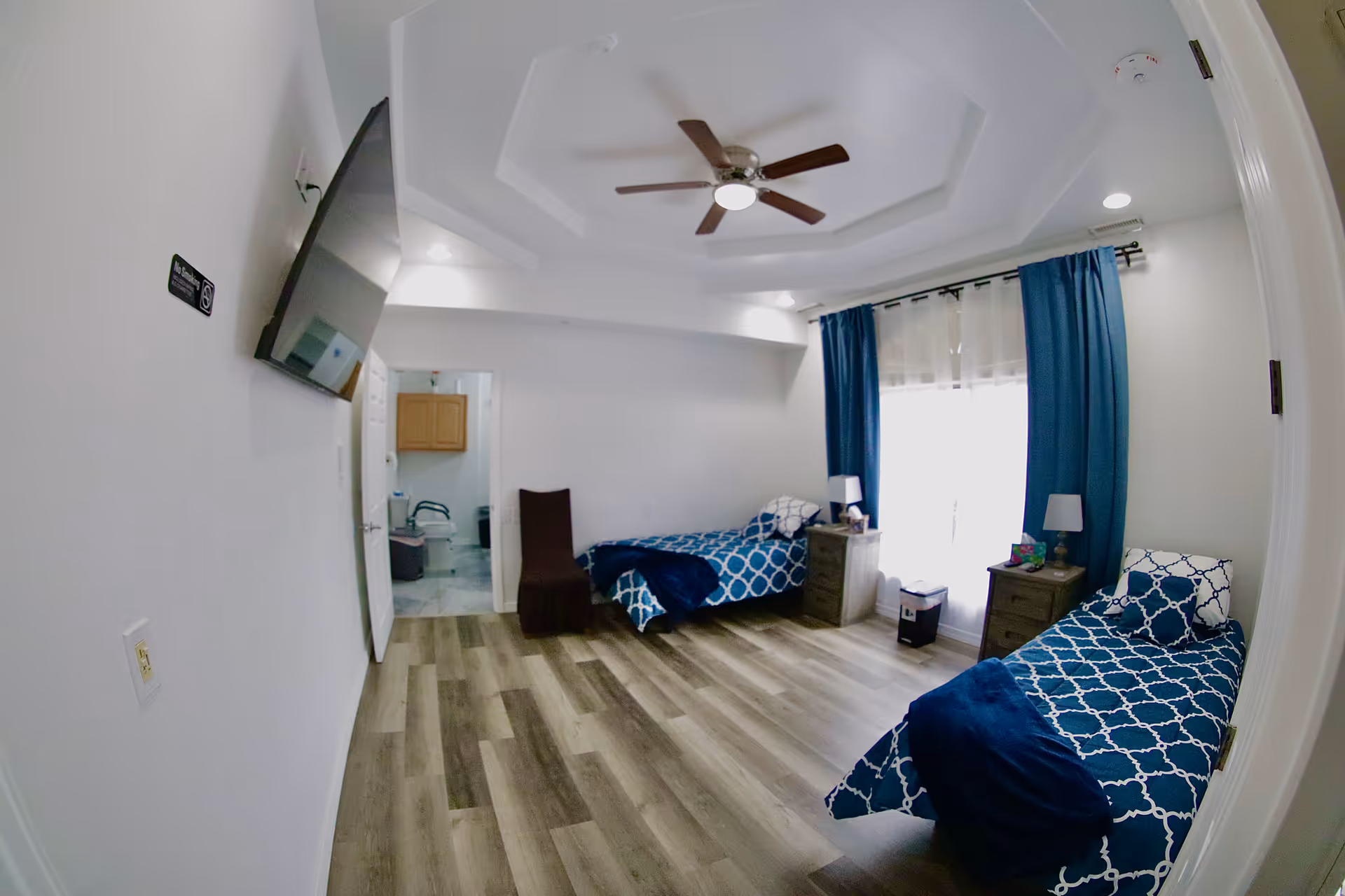 Two blue patterned beds in a bright shared senior living bedroom with ceiling fan and curtains.