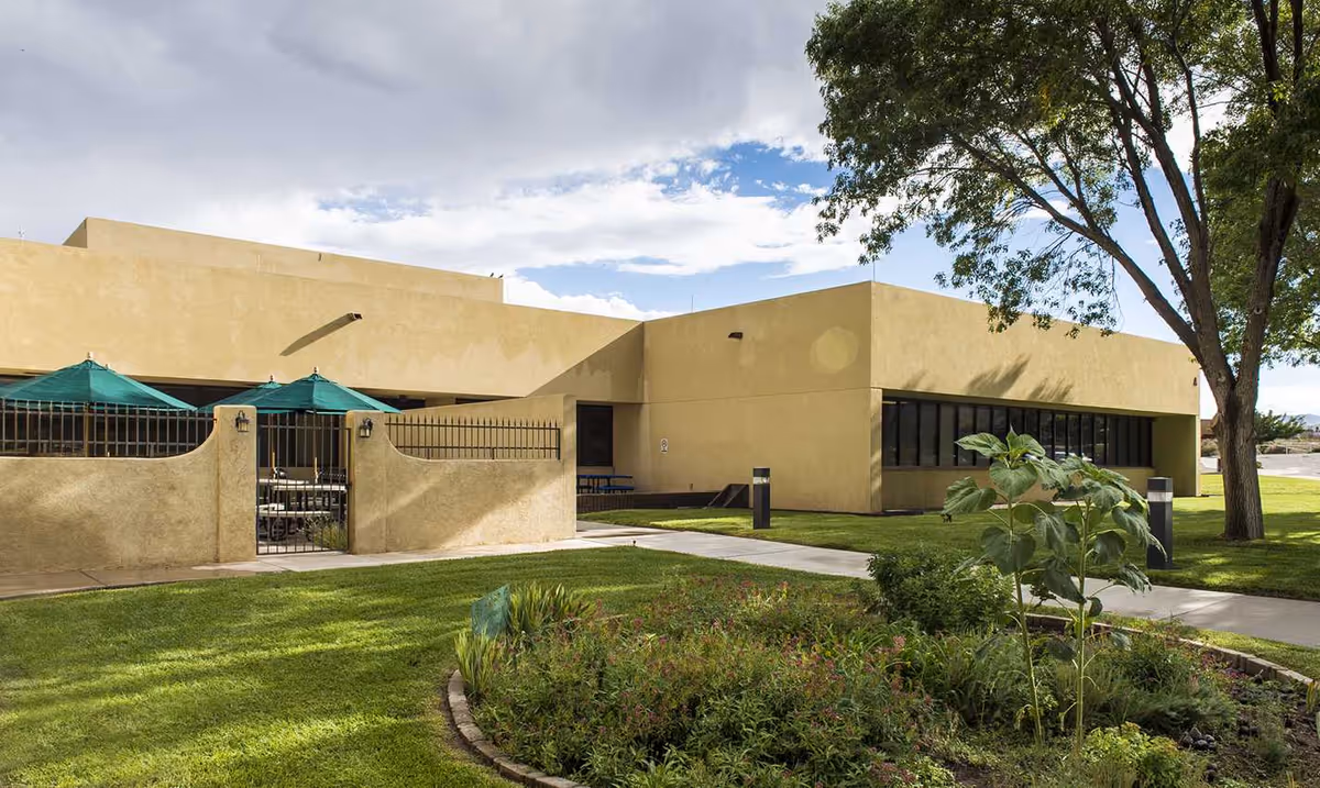 Exterior view of a beige building with a green lawn, trees, and outdoor seating.