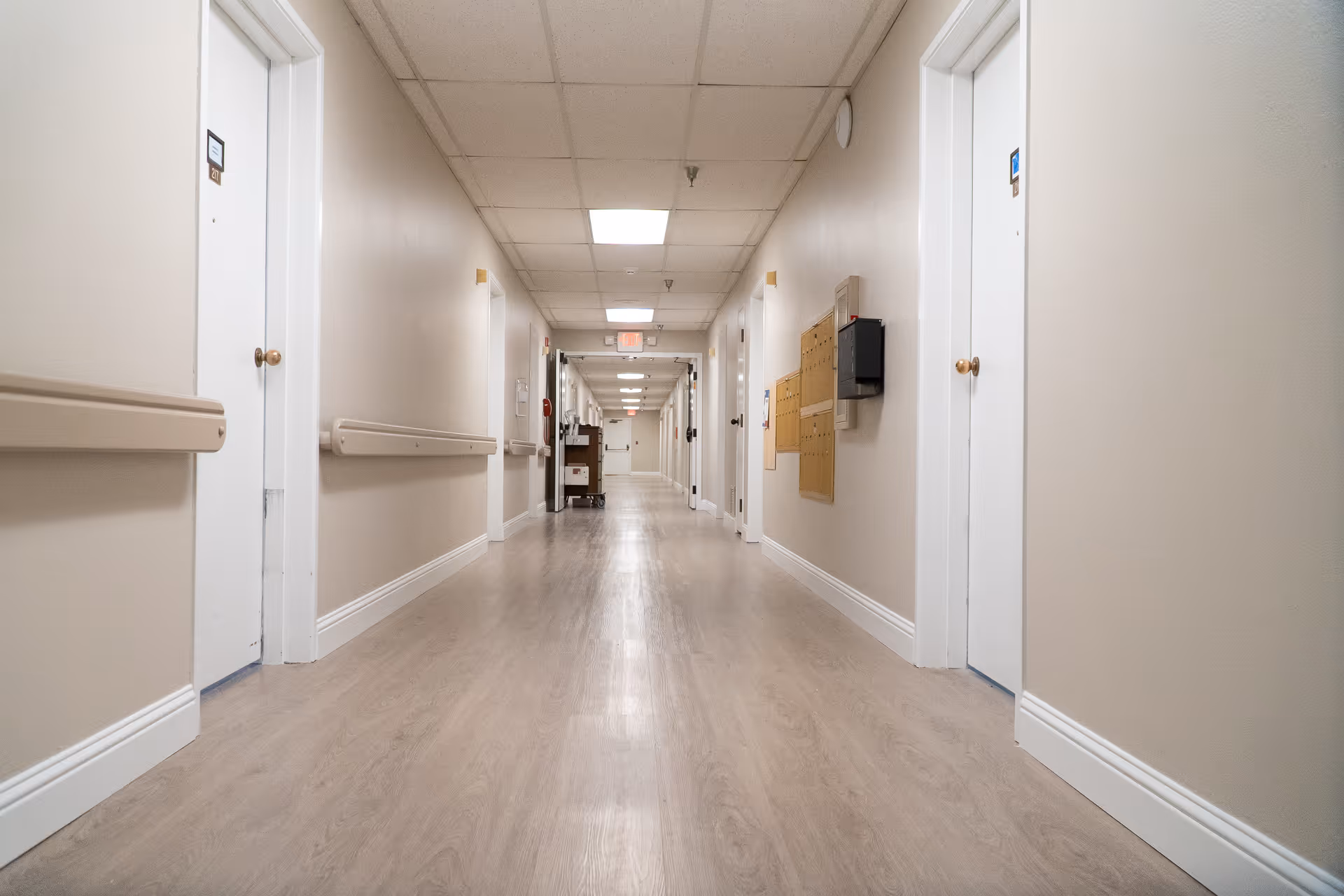 A long, clean hallway in a senior living facility with beige walls, light wood flooring, white doors on both sides, handrails along the walls, and ceiling lights illuminating the corridor.