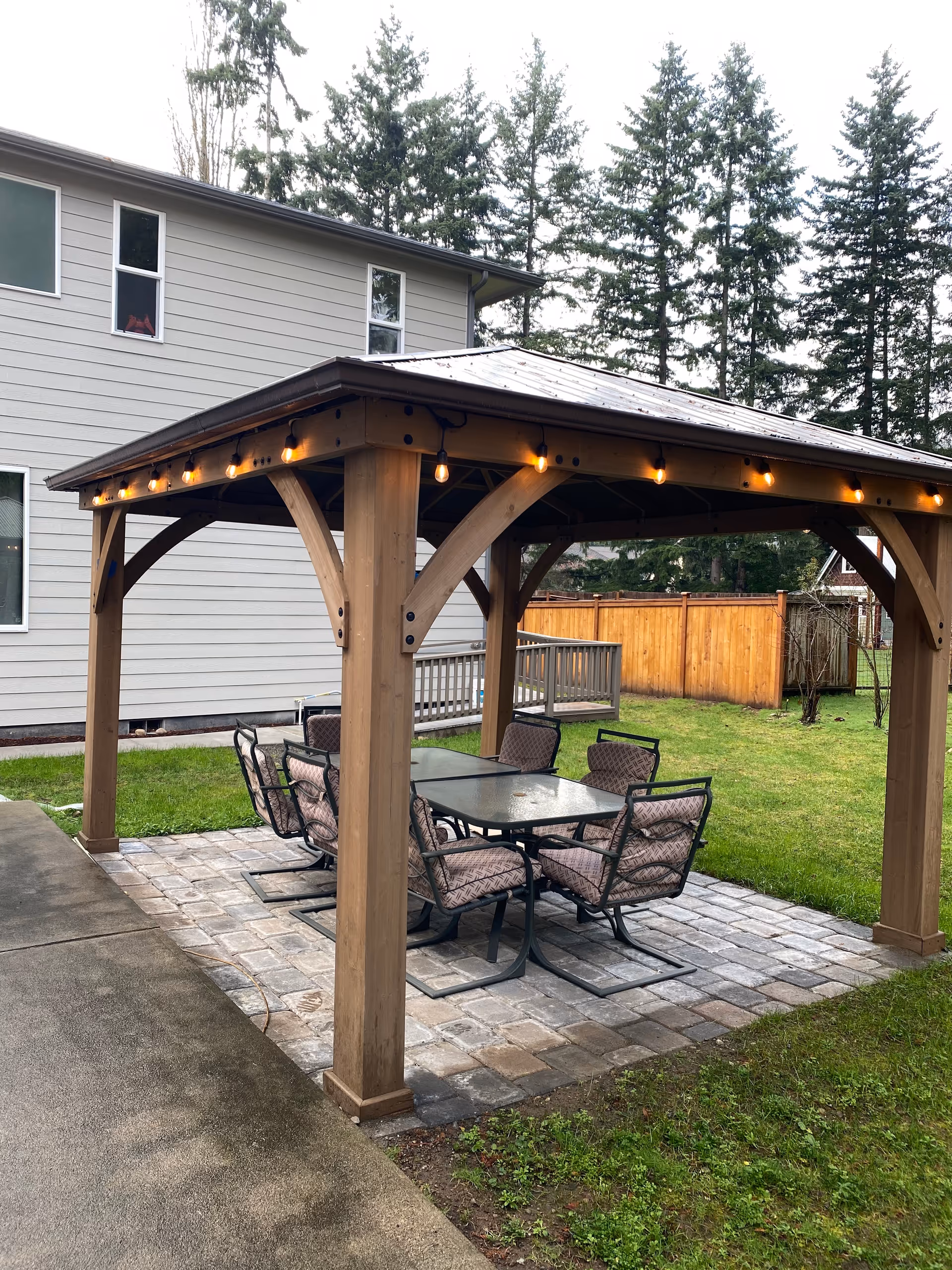 Wooden gazebo with string lights and a glass-table dining set in a backyard.