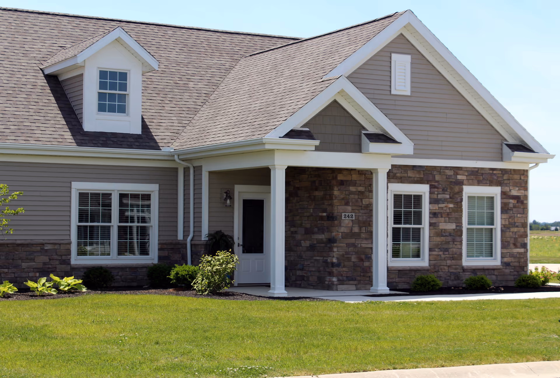 Exterior view of a single-story residential building with beige siding and stone accents, featuring a small covered porch with white columns, multiple windows with white trim, and a well-maintained green lawn in front.