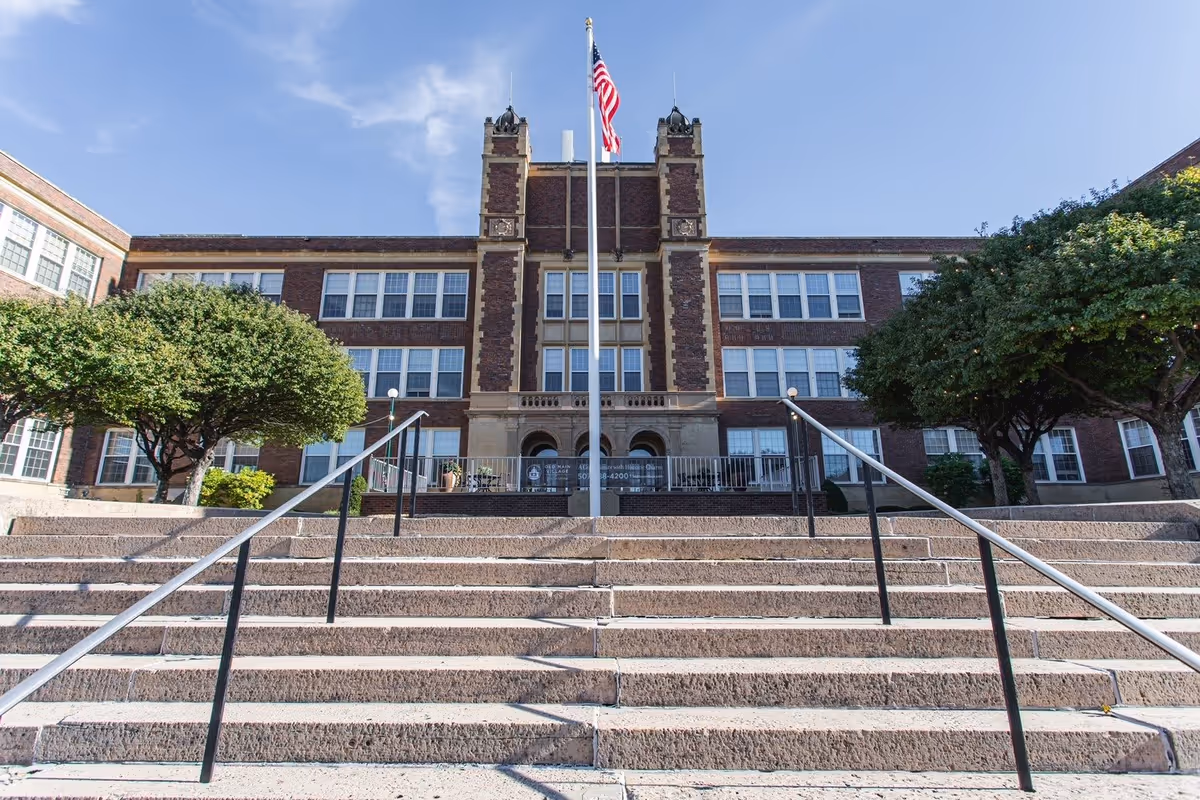 Front view of a symmetrical brick building with wide stone steps, handrails, an American flagpole, and trees on either side.