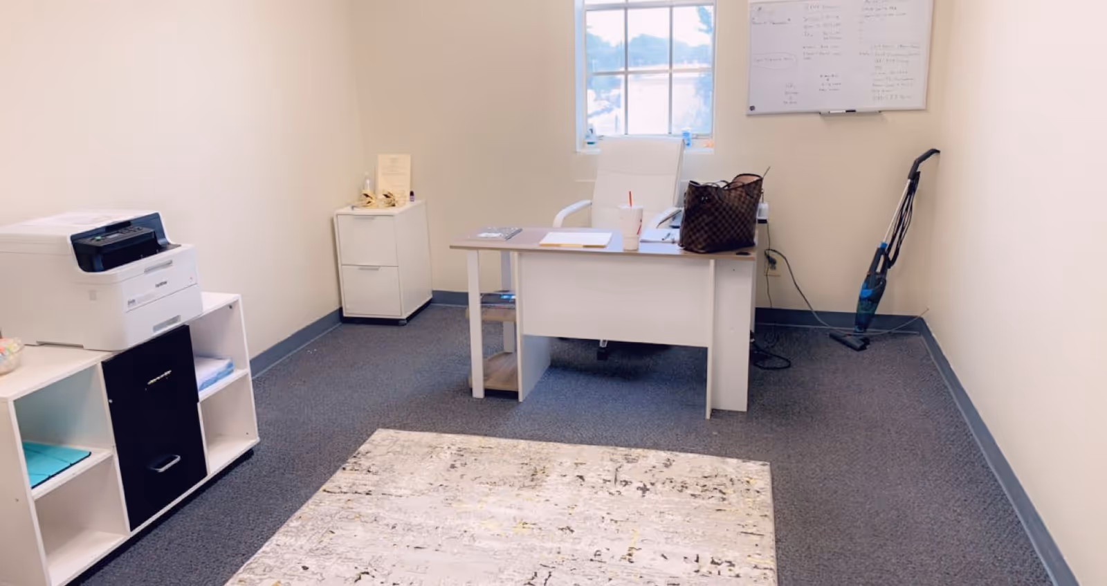 An office room with desk, printer, shelves, and a vacuum.