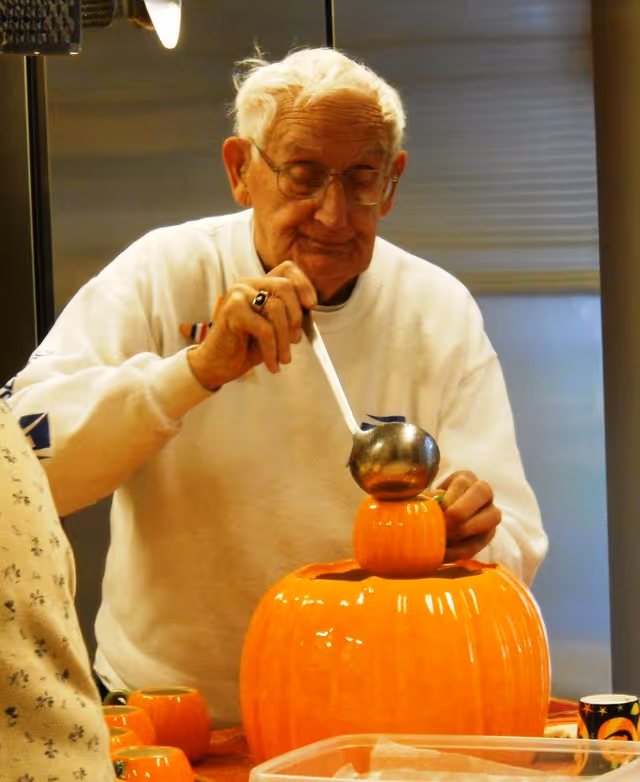 An elderly man wearing glasses and a white sweatshirt is ladling a beverage from a large orange pumpkin-shaped container into a smaller matching pumpkin-shaped cup. The setting appears to be a kitchen with a refrigerator in the background and other pumpkin-themed cups on the table.