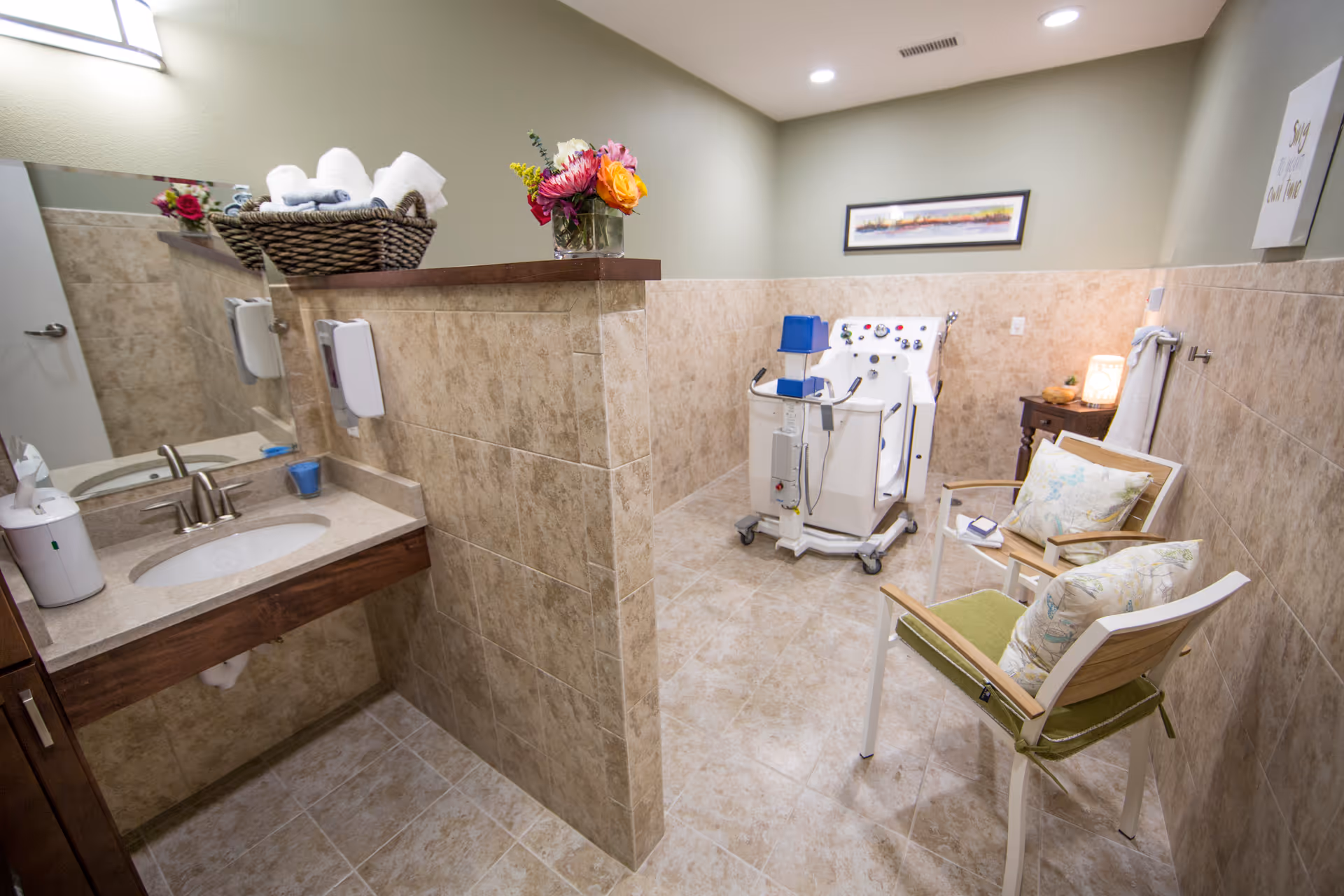 Tiled facility bathroom with sink, mirror, towels, and a waiting chair beside medical equipment