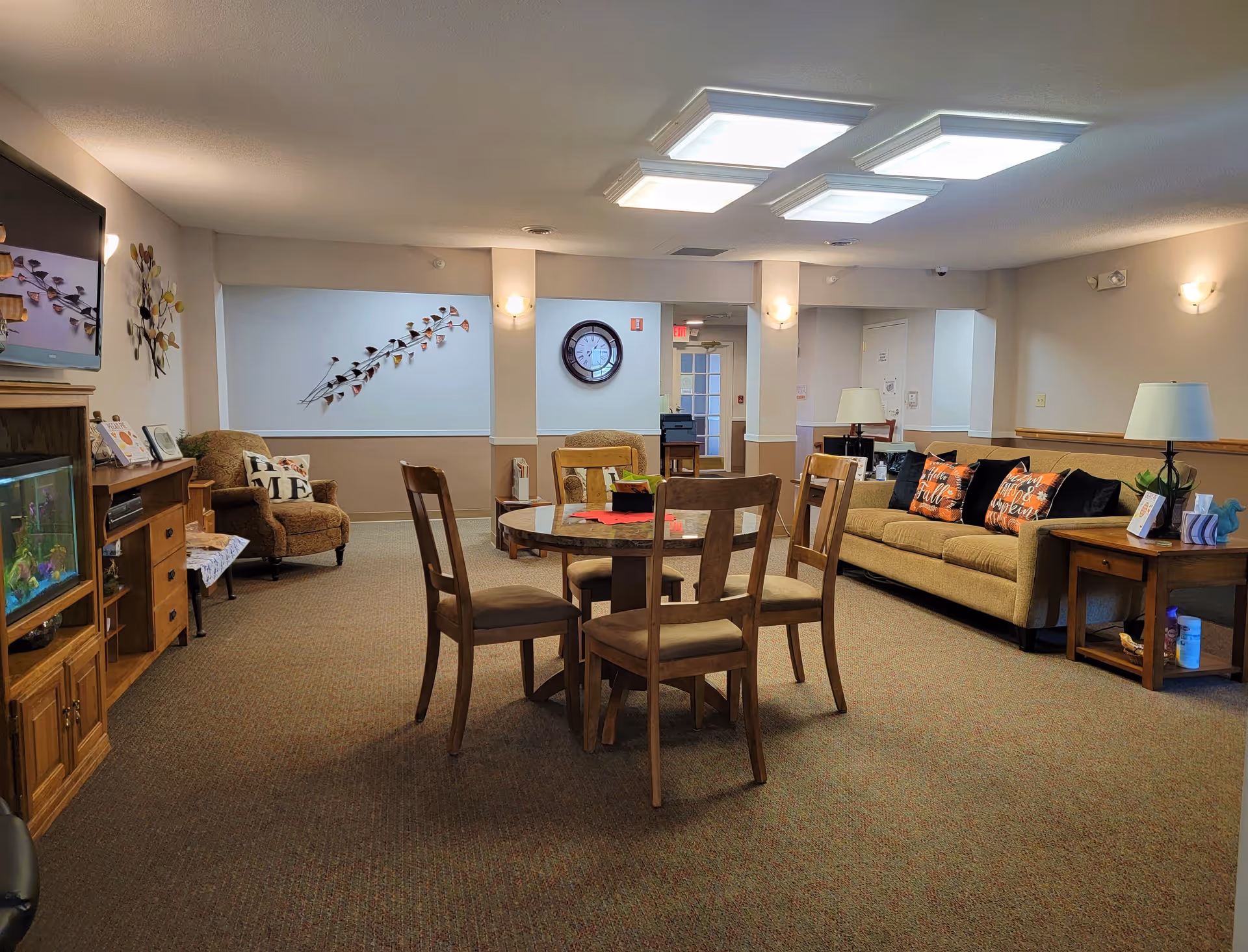 A cozy senior living common area with a round wooden table and four chairs in the center. To the right is a beige sofa with three decorative pillows featuring fall-themed designs. There are two armchairs, one on each side of the room, and a wooden cabinet with a fish tank on the left. The walls are decorated with metal wall art and a clock. The room is well-lit with ceiling lights and wall sconces.