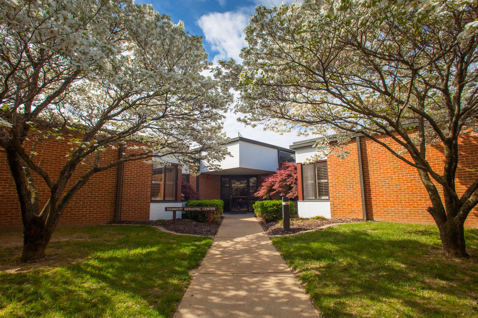 Exterior view of the Benedictine Living Community-At The Shrine showing a brick building with a pathway leading to the entrance. The entrance is framed by two trees with white blossoms and some bushes near the doorway. A sign near the entrance reads 'Dammert Geriatric Center'.