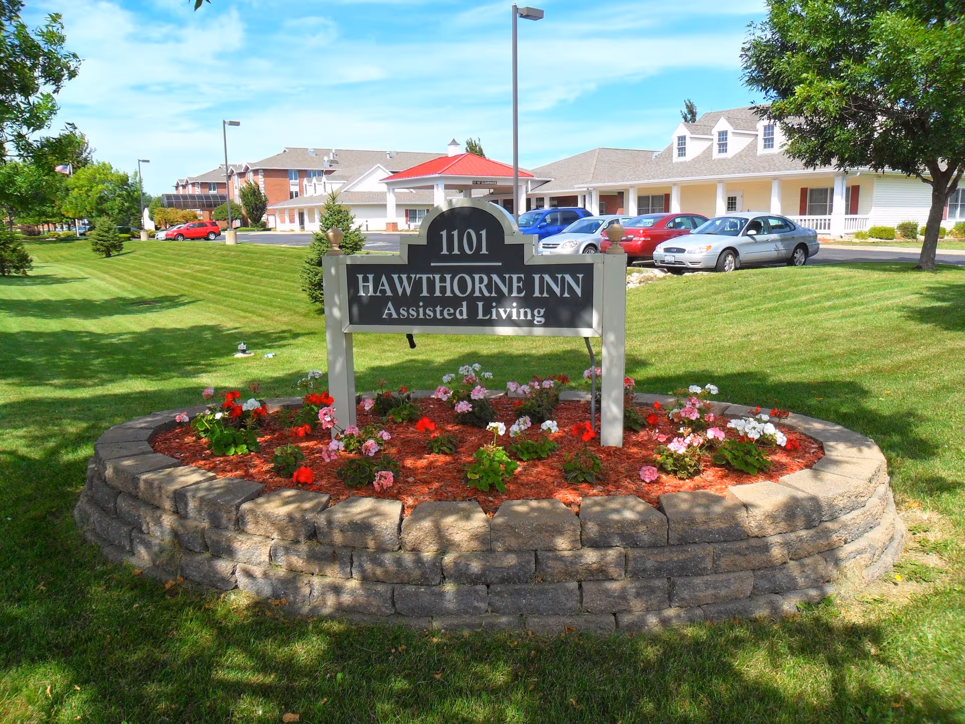 A landscaped flower bed with red and pink flowers surrounds a sign that reads '1101 Hawthorne Inn Assisted Living'. Behind the sign is a parking lot with several cars and a single-story building with a red roof and white exterior. The sky is blue with some clouds, and there are trees and well-maintained grass around the area.
