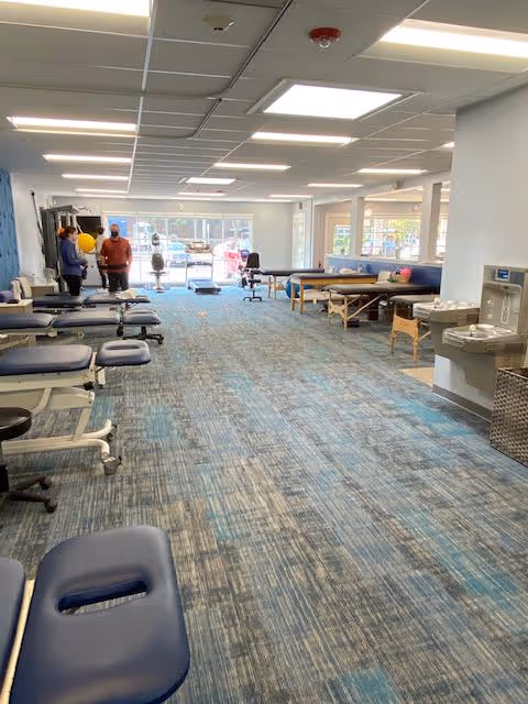 Wide therapy room with multiple treatment tables, chairs, and staff visible in distance.
