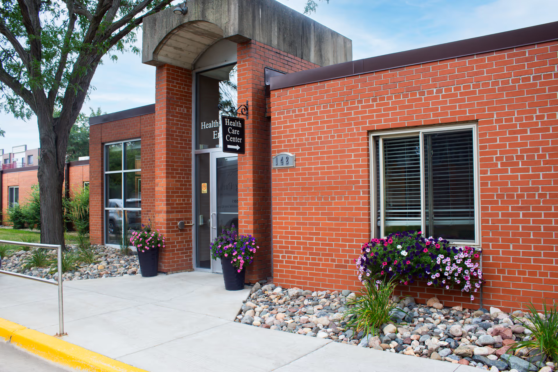 Red brick senior living entrance with planters, rocks along walkway, and a glass door