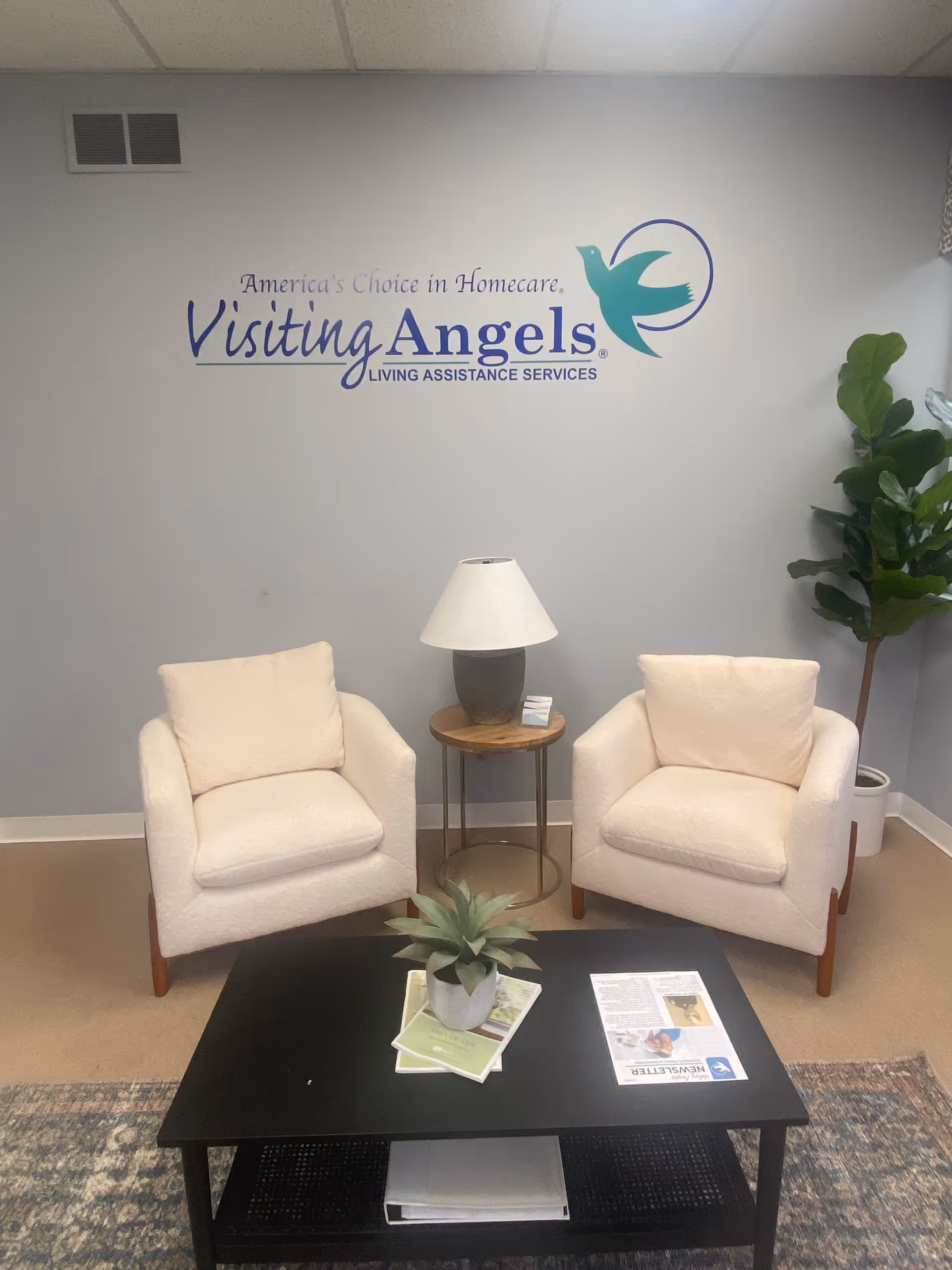 Two white armchairs flank a coffee table in a lounge beneath the Visiting Angels logo.