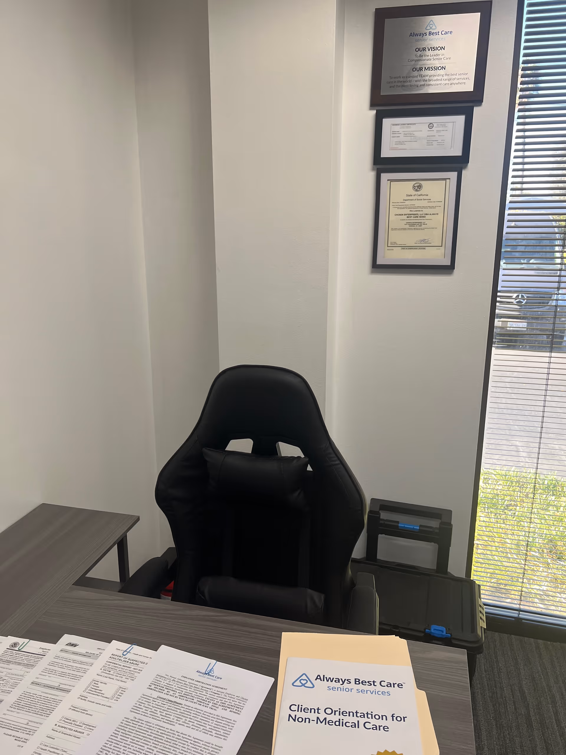 An office desk with a black chair, papers, and framed certificates on the wall.