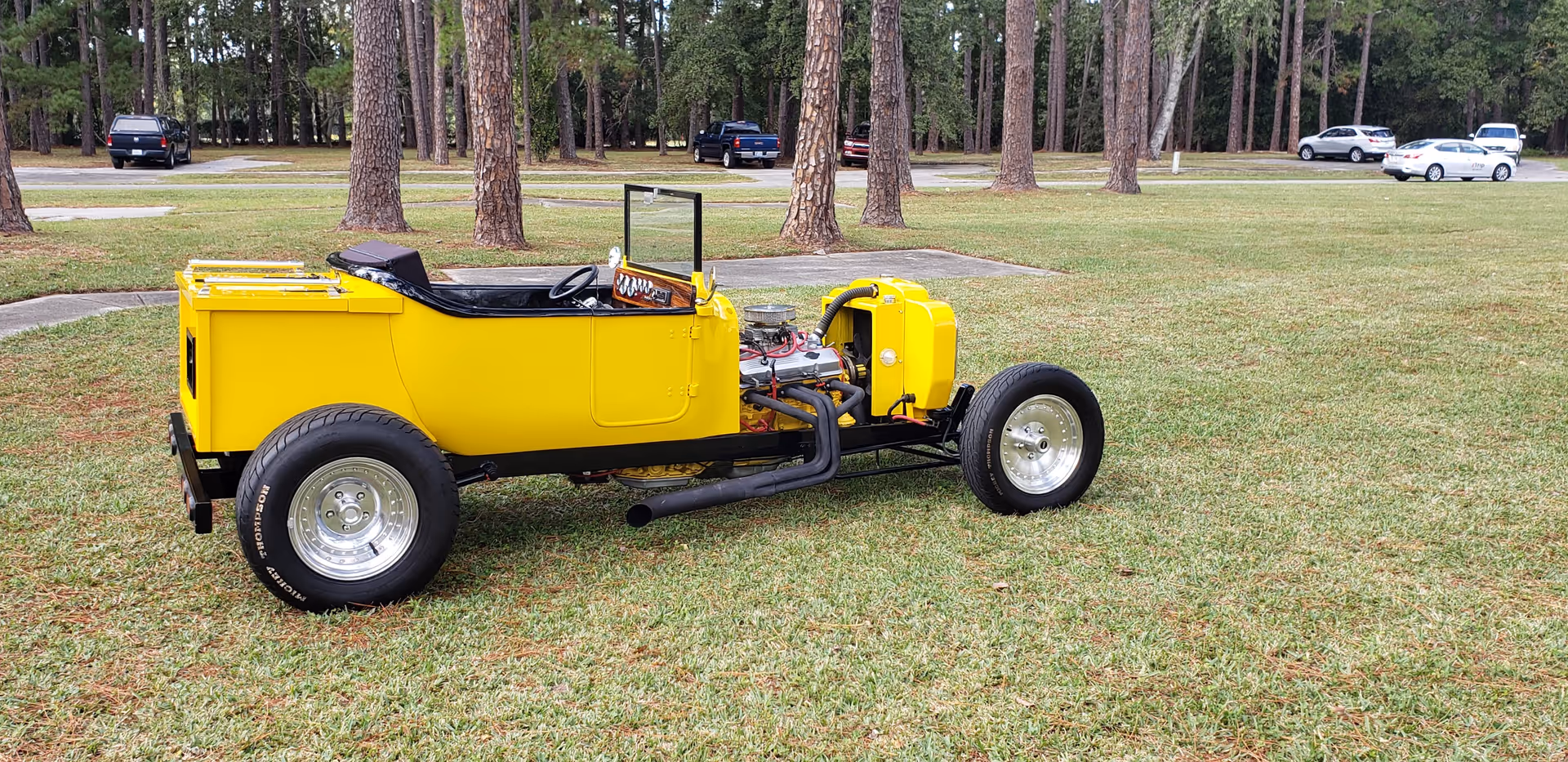 A bright yellow vintage convertible parked on a grassy lawn among tall trees.