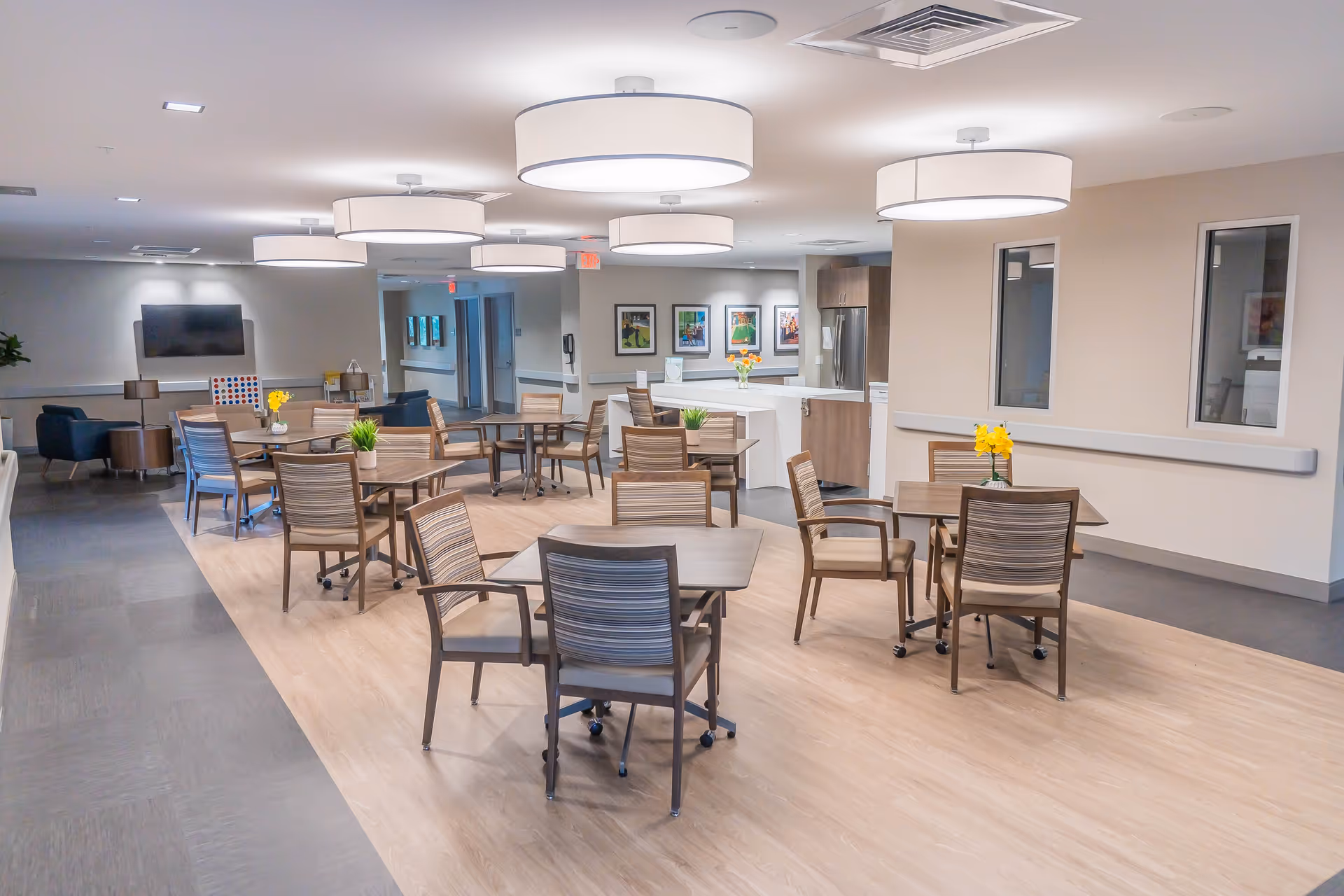A bright and modern dining area in a senior living facility with several wooden tables and chairs arranged neatly. The room features large circular ceiling lights, a small kitchen area with a refrigerator and counter, framed artwork on the walls, and a television mounted on the far wall. There are small plants and flowers on some tables, and a seating area with armchairs is visible in the background.