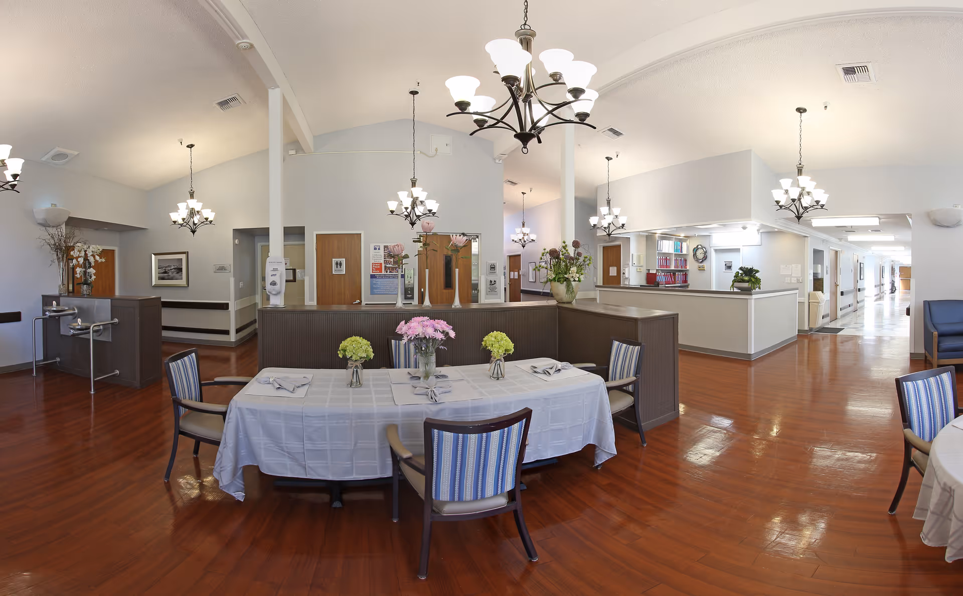 Interior view of a senior living facility dining area with a table covered in a white tablecloth, set with napkins and flower vases. The room has wooden floors, multiple chandeliers, and a reception or nurse station in the background. Hallways and doors are visible leading to other parts of the facility.