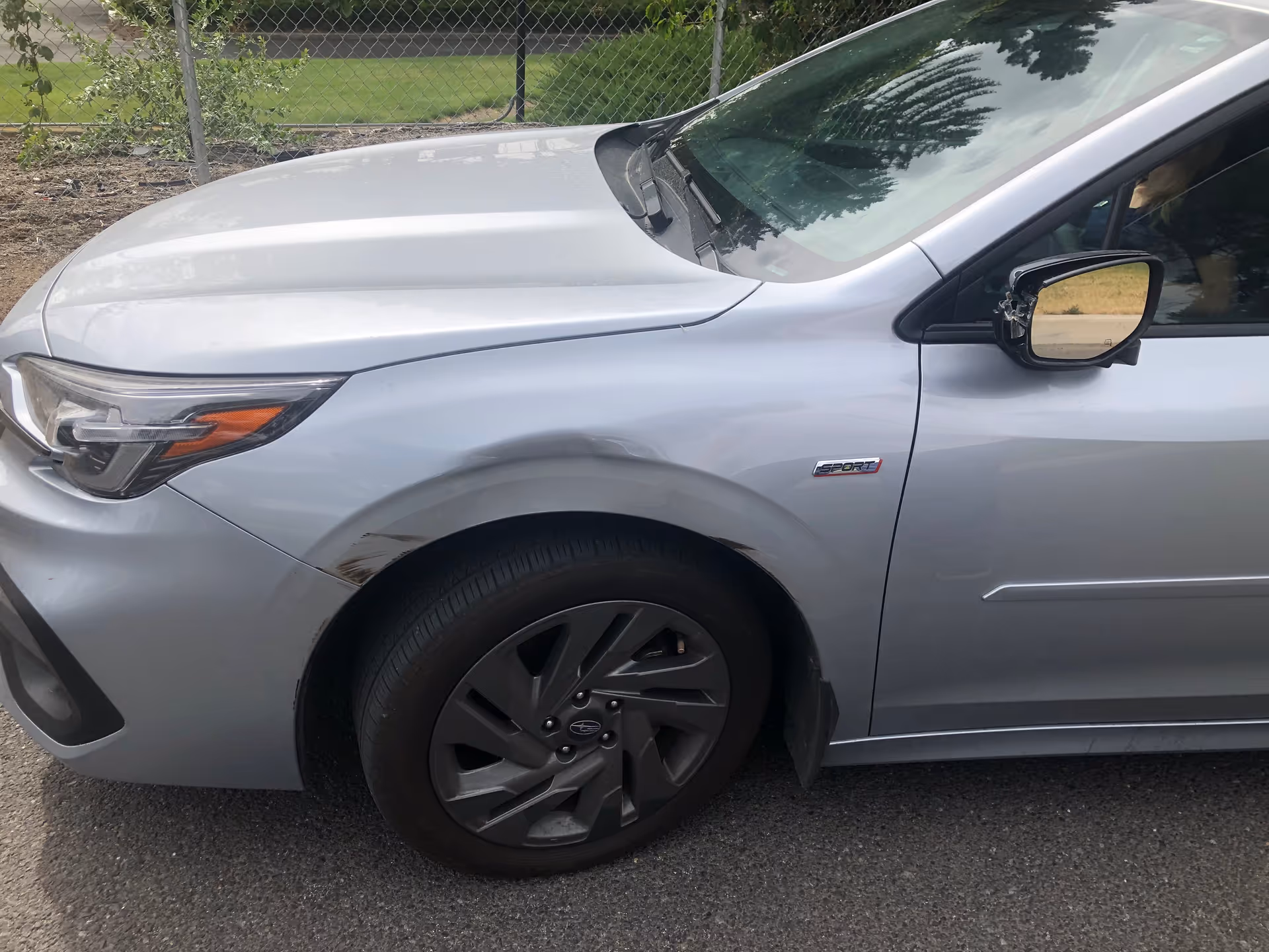 Close-up side view of a silver car with a dented fender outdoors near a fence.