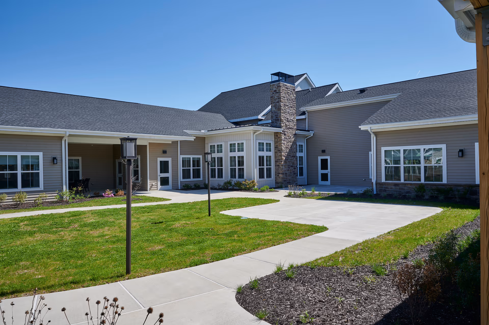 Courtyard and paved walkways in front of a single-story senior living building with large windows and a stone chimney.