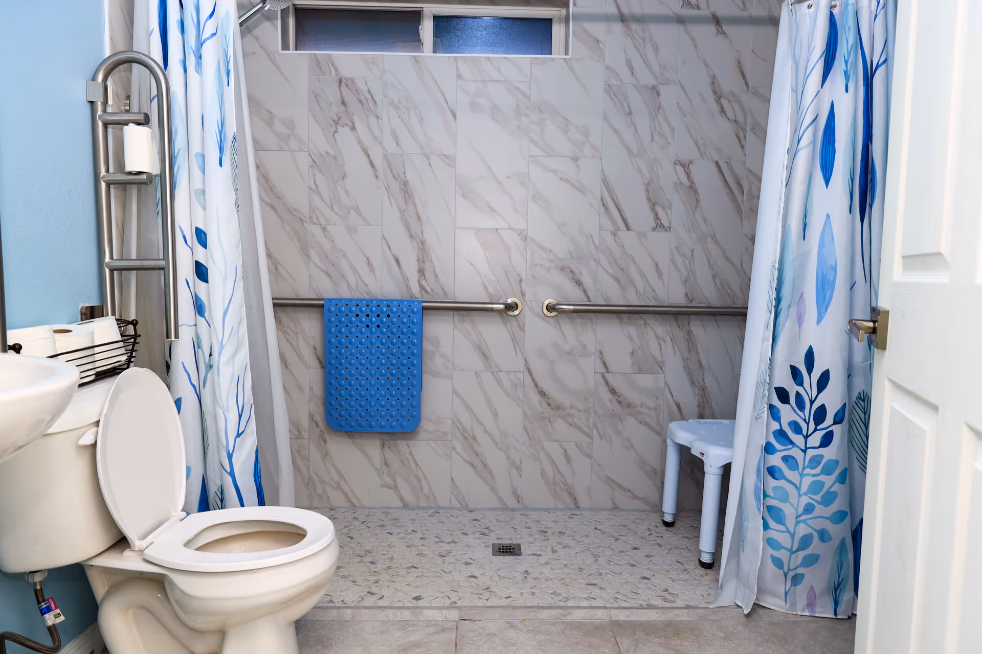 Accessible bathroom with toilet, shower area, grab bars, blue mat, and patterned shower curtains.