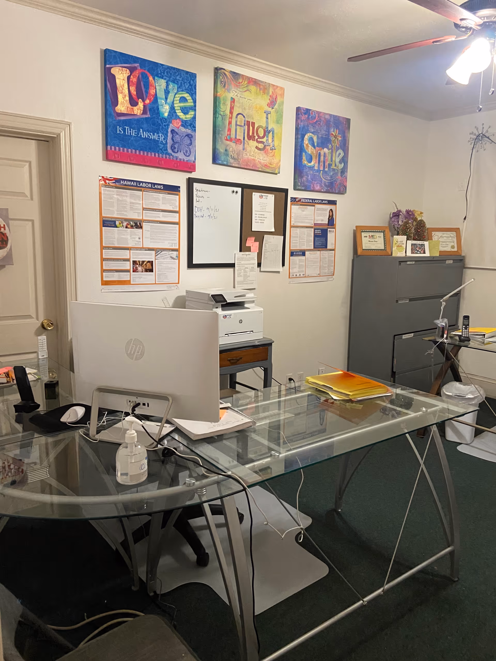 Glass desk office with computer, printer, filing cabinet, and colorful wall art.
