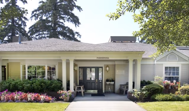 Single-story building with a covered entrance supported by columns, surrounded by green shrubs and colorful flowers, with trees in the background under a clear sky.