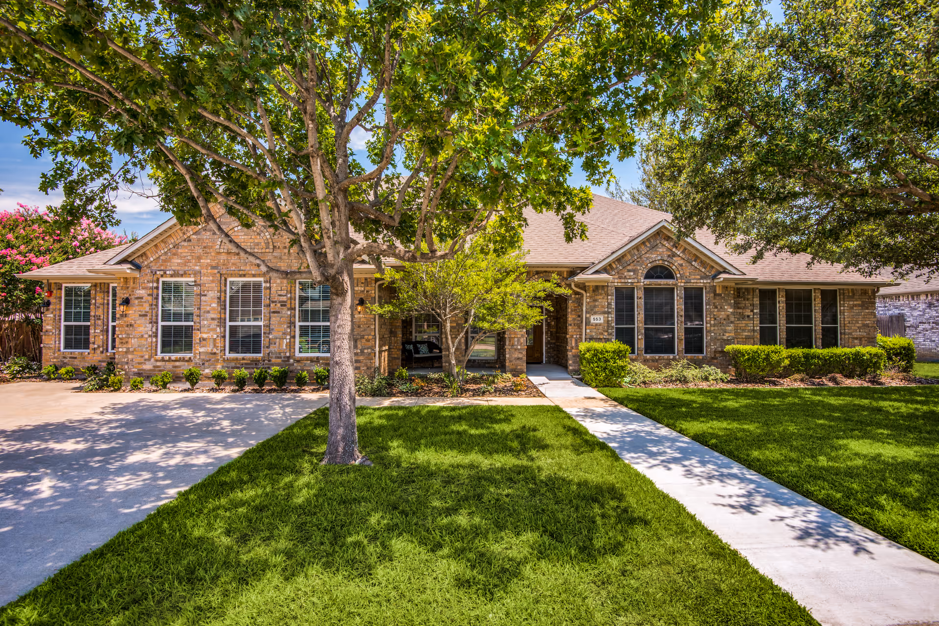 Brick assisted living facility with a manicured lawn, trees, and a concrete walkway to the entrance.