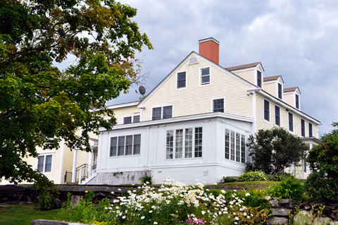 Two-story beige senior living home with a sun porch, surrounded by trees and blooming garden.