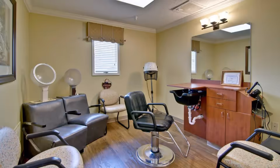 Interior of a hair salon area in a senior living facility with a black salon chair in front of a wooden counter with a sink, a large mirror, and hair styling equipment. There are additional chairs along the walls and a window with blinds.