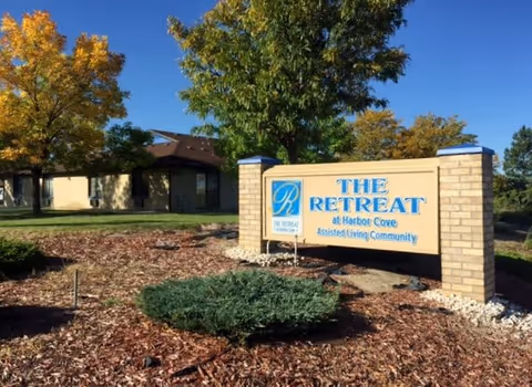 Outdoor view of the entrance sign for The Retreat at Harbor Cove Assisted Living Community, surrounded by landscaping with trees and shrubs under a clear blue sky.