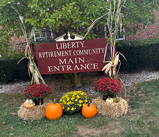 Wooden sign reading "Liberty Retirement Community Main Entrance" decorated with pumpkins, hay bales and fall flowers in front of a brick building.