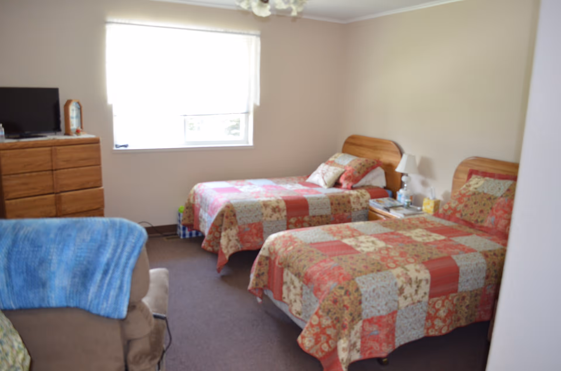 A bedroom with two twin beds covered in patchwork quilts in shades of red, beige, and green. There is a wooden dresser with a TV on top against the left wall, a window with a white blind letting in natural light, and a beige armchair with a blue blanket draped over it in the foreground.