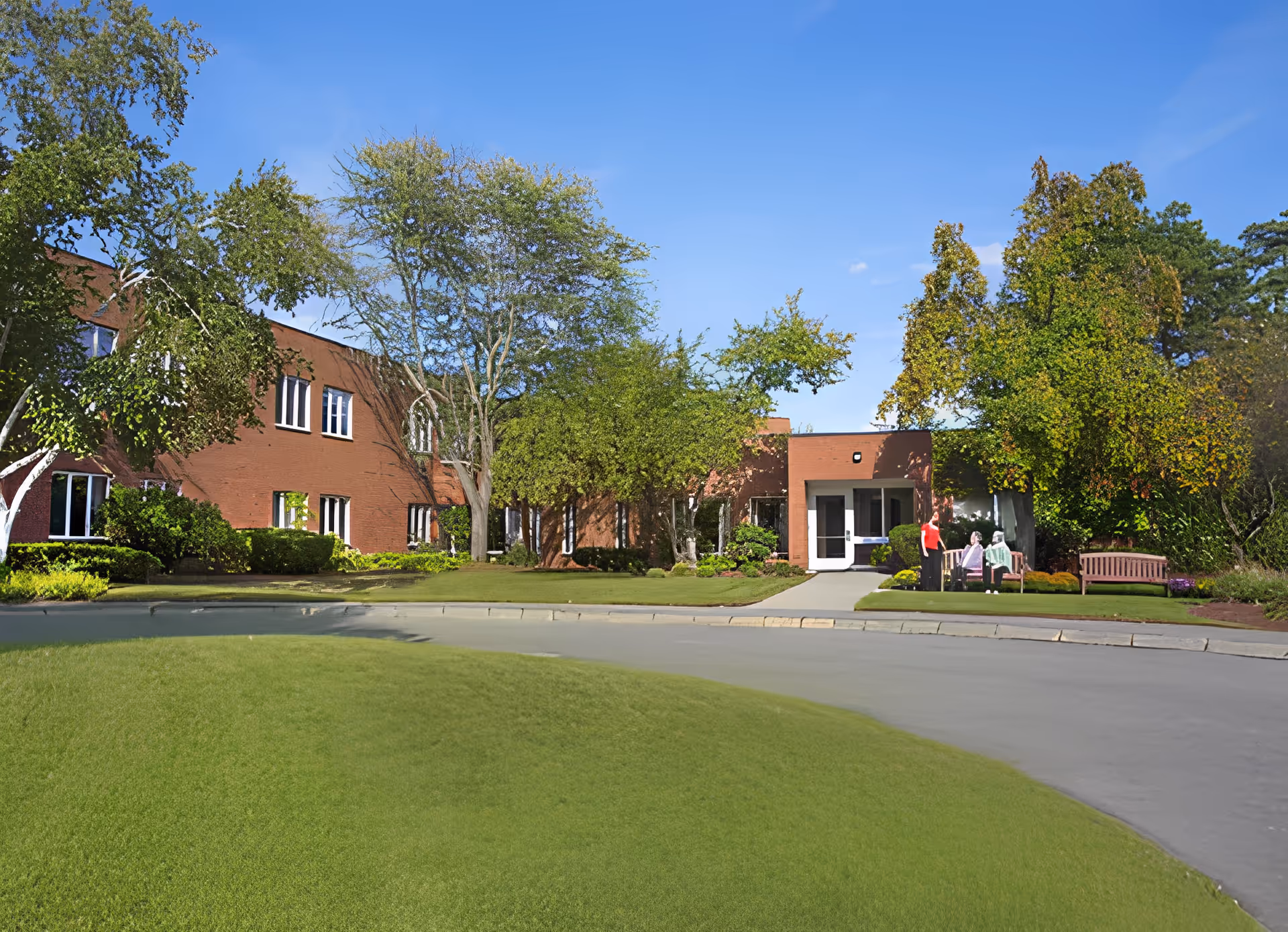 Front view of a two-story brick senior living building with a grassy lawn, trees, walkway and benches with a few people near the entrance.