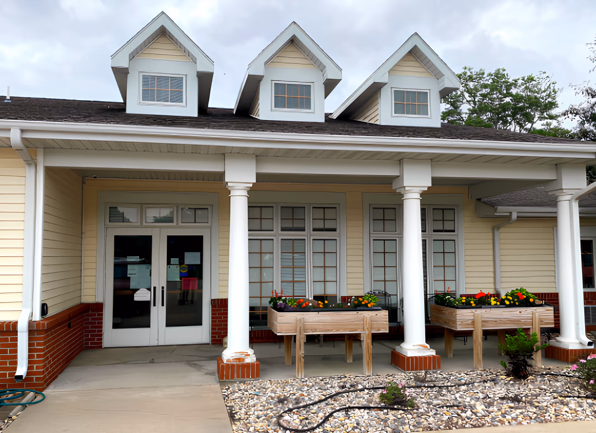 Exterior view of a building with a covered porch supported by white columns. The building has beige siding with red brick accents and three dormer windows on the roof. There are two raised wooden garden beds with colorful flowers on the porch, and a concrete walkway leading to double glass doors.