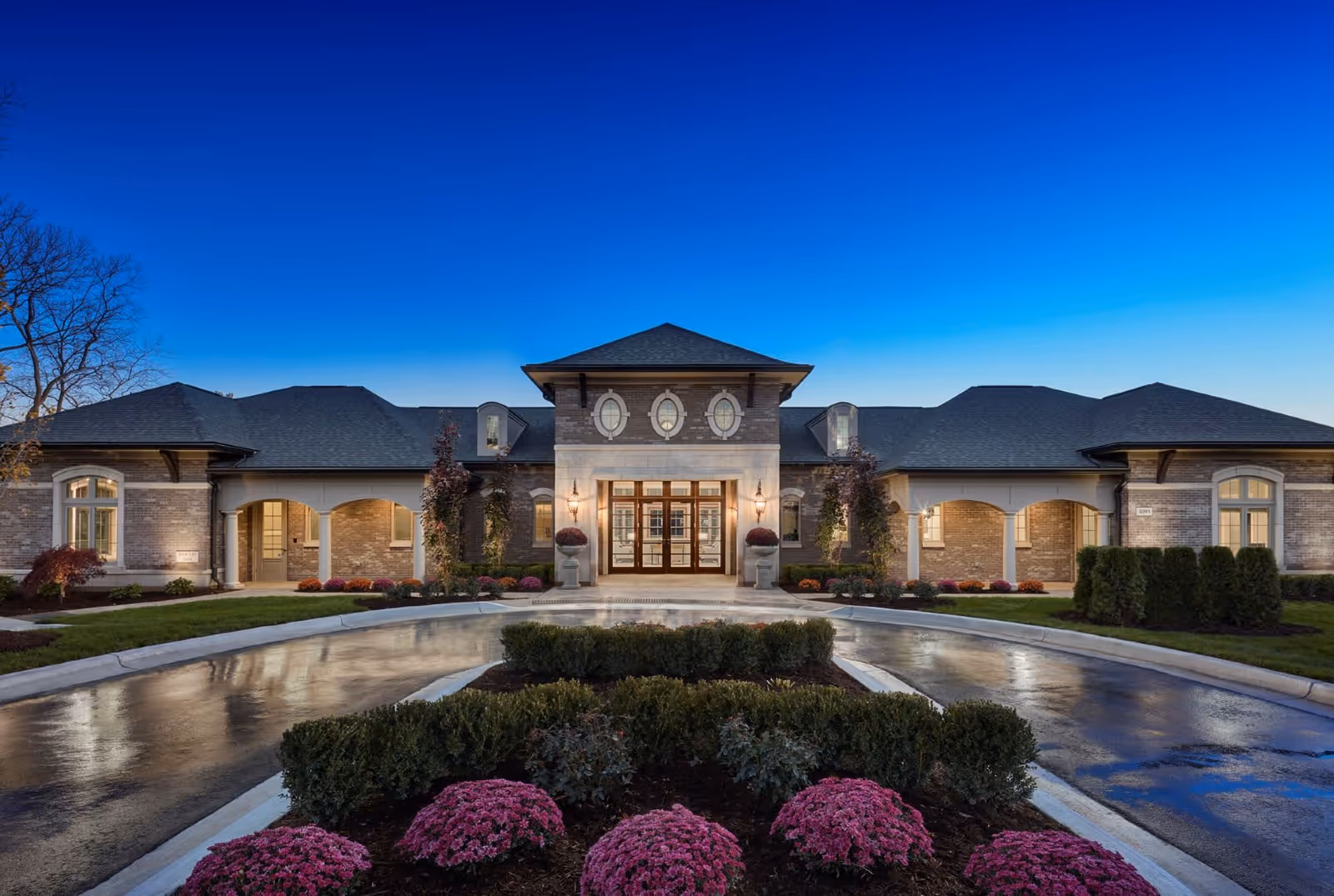 Front exterior view of a large, elegant senior living facility building at dusk with well-maintained landscaping, including bushes and pink flowers, and a circular driveway leading to the main entrance with glass doors and decorative windows above.