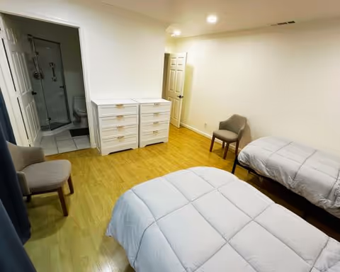 A bright bedroom with two single beds, a dresser, chairs, and an ensuite bathroom.