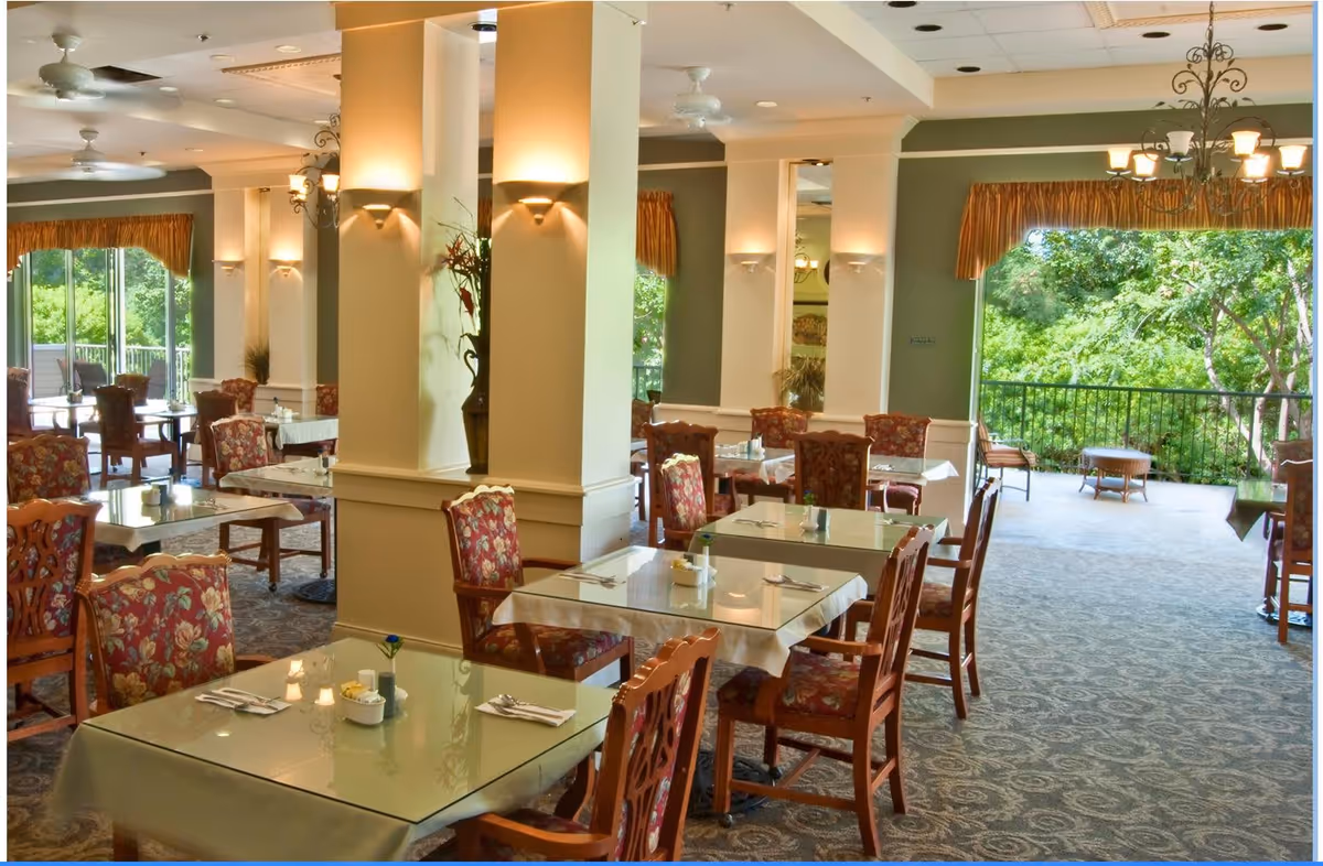 Bright dining room with glass-topped tables and floral-upholstered chairs opening onto a leafy outdoor patio.