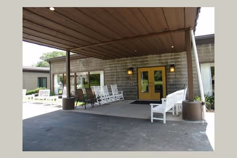 Covered entrance area of a building with multiple white rocking chairs and a few brown chairs arranged along the walls. The building has double yellow doors and large windows, with a brown roof overhang providing shade.